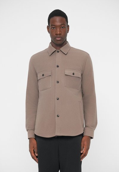 Veste en chemise marron à boutons avec deux poches poitrine, boutons noirs, col, et un tissu lisse et texturé. Design ajusté.