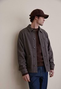 Brun uld skjortejakke med krave, frontknapper og sidelommer. Båret over en brun sweater og parret med blå jeans.