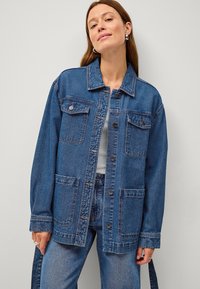 Giacca in denim, blu scuro, vestibilità rilassata, con tasche frontali, chiusura a bottoni e colletto classico. Tessuto testurizzato con cuciture visibili.