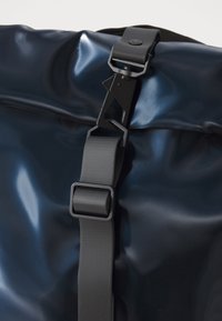 Close-up of a dark blue, glossy backpack strap and buckle, highlighting the sleek material and functional design details.