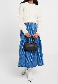 Sweter w kolorze kremowym z dzianiny, niebieska spódnica maxi w wzory, czarna cylindryczna torebka i białe botki za kostkę. Skoncentruj się na fakturach i kontrastach kolorystycznych.