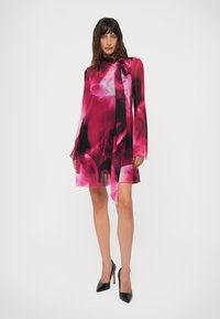 Mulher vestindo um vestido de mangas longas, com estampado floral em rosa e preto, com um laço no pescoço, combinado com sapatos de salto alto pretos, de pé contra um fundo branco.