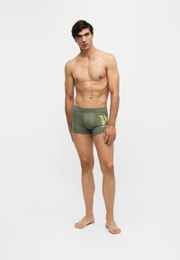 Homme torse nu debout pieds nus, portant un boxer vert avec un motif jaune sur la cuisse gauche, sur fond blanc uni.