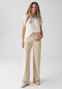 Beige Corduroy-Hosen mit weitem Bein kombiniert mit einem cremefarbenen Strickoberteil und einem hellbraunen Schal, die einen entspannten Schnitt und weiche Texturen zeigen.