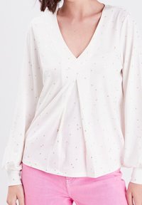 Weiße Bluse mit tiefem V-Ausschnitt, langen Ärmeln, verziert mit goldenen Sprenkeln und Plissee-Details. Kombiniert mit rosa Hosen für einen Kontrast.