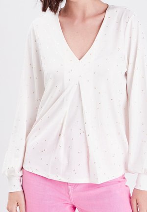 Weiße Bluse mit tiefem V-Ausschnitt, langen Ärmeln, verziert mit goldenen Sprenkeln und Plissee-Details. Kombiniert mit rosa Hosen für einen Kontrast.