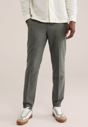 Man draagt grijze op maat gemaakte broek, witte en beige sneakers en een crèmekleurig overhemd met knopen, met de handen gedeeltelijk in de zakken.