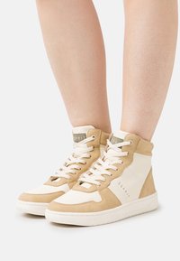 Beige und cremefarbene High-Top-Sneaker aus verschiedenen Materialien, mit strukturierter Oberfläche, Schnürung vorne und Logoakzent auf der Zunge. Flache Gummisohle.