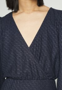 Vestido envelopado azul escuro, com um tecido texturizado e um decote em V profundo, além de detalhes franzidos na cintura. Mangas longas bufantes.