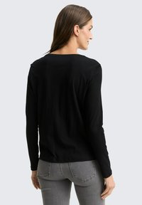 Woman with long brown hair wearing a black long-sleeve top and gray jeans, facing away against a plain light background.