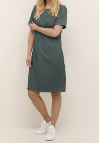 Vestido de teal oscuro hasta la rodilla con mangas cortas, escote redondo y un bolsillo lateral. Combinado con zapatillas blancas. Textura de tela suave.