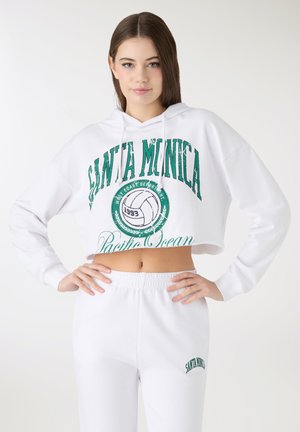 Felpa bianca corta con stampa grafica verde e coulisse, abbinata a pantaloni della tuta bianchi con testo verde coordinato.