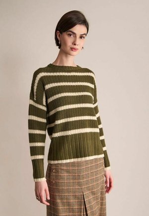 Femme portant un pull en maille rayé vert olive et blanc et une jupe marron à carreaux, debout devant un fond beige uni.