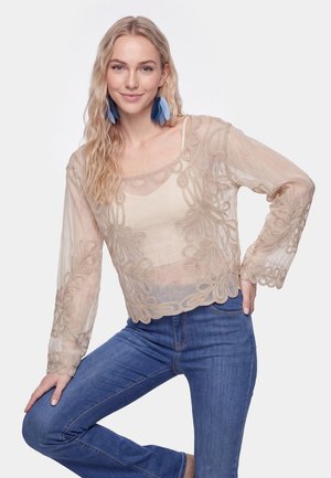 Vrouw met lang blond haar die blauwe veeroorbellen draagt, een doorschijnende beige geborduurde top over een beige hemdje en blauwe spijkerbroek, poserend met één opgeheven been.