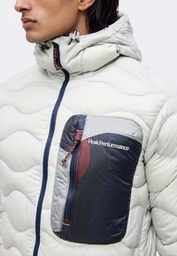 Gros plan d'une personne portant une veste matelassée gris clair avec capuche et une poche poitrine multicolore avec fermeture éclair et le logo "Peak Performance".