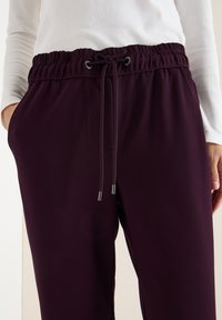 Fialové jogger kalhoty s elastickým pasem, stahovací šňůrkou a postranními kapsami. Hladká textura, přiléhavý střih pro pohodlí.