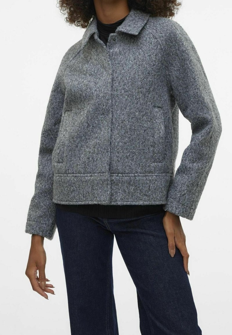 Femme portant une veste en laine grise texturée avec col, poches avant et jean bleu foncé, posant avec une main sur la hanche.
