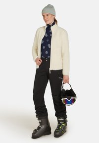 Fleecejacke in Creme, marineblaue gemusterte Unterziehshirt, schwarze Skihosen und schwarze Skistiefel. Hält einen Helm mit reflektierendem Visier.