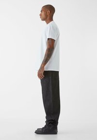T-shirt bianco a maniche corte abbinato a pantaloni in denim scuro, larghi, e sneakers nere. La vista laterale mette in evidenza la vestibilità e lo stile.