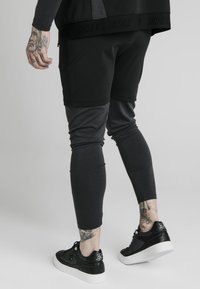 Black and gray athletic pants with fitted legs, featuring a textured waistband and logo. Worn with black sneakers and visible tattoos.