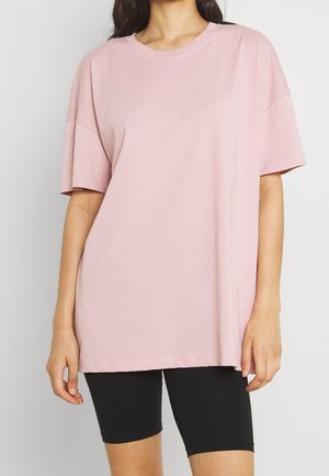 Kvinde iført en løs lyserød kortærmet t-shirt og stramme sorte cykelshorts mod en ensfarvet baggrund.