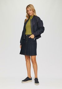 Donkerdenim bomberjack met geribbelde manchetten, gecombineerd met een strakke groene top en een flared denimrok, verfraaid met zichtbare stiksels. Zwarte sneakers.