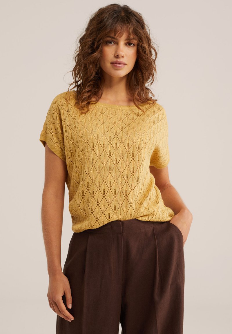 Sieviete ar cirtainiem brūniem matiem, tērpusies dzeltenā teksturētā īsām piedurknēm topā un tumši brūnās biksēs ar augstu jostasvietu, ar vienu roku kabatā.