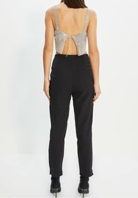 Zadní pohled na ženu v metalickém halter topu s vzorem a mašlí, doplněném o černé kalhoty s vysokým pasem a kotníkovými botami.