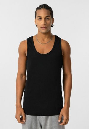 Zwarte geribbelde tanktop met brede schouderbanden. Heeft een ronde halslijn en een aansluitende pasvorm. Gemaakt in combinatie met grijze shorts, op een effen achtergrond.