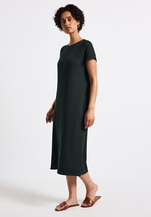 Abito a coste verde scuro con maniche corte, che arriva fino a metà polpaccio. Indossato con sandali marroni a infradito. Tessuto liscio e silhouette semplice.