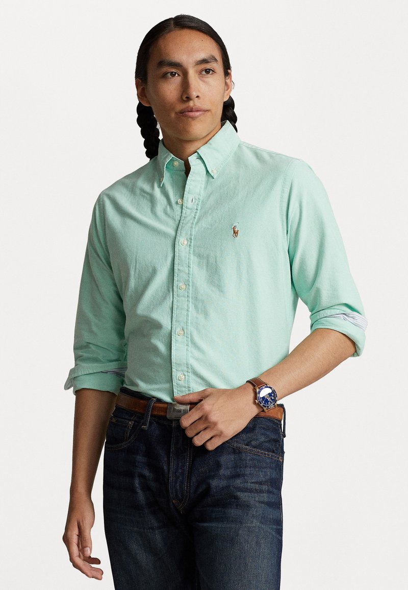 Light green button-up shirt with long sleeves rolled at cuffs, small logo on chest, paired with dark blue denim jeans and a watch.