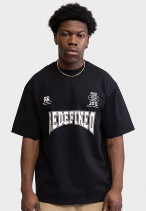 Jeune homme aux cheveux courts et bouclés porte un t-shirt noir oversized avec le texte blanc "REDEFINED" et un pantalon beige sur fond gris uni.
