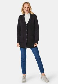 Dunkelgrauer Cardigan mit Schalkragen, ausgestattet mit fünf Knöpfen, zwei vorderen Taschen und einem strukturierten Strickmuster. Kombiniert mit blauen Jeans und hellen Sneakers.