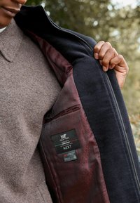 Giacca della marina con fodera in tessuto grigio-marrone, dotata di un interno a motivi, chiusura con zip e etichette visibili sulla tasca interna.