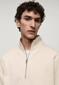 Beige Kapuzenpullover mit Reißverschluss, der einen hohen Kragen, eine weiche Textur und einen silbernen Reißverschluss aufweist, über einem weißen T-Shirt getragen.