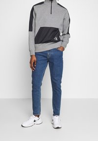 Felpa grigia con zip a metà, pannello nero e dettaglio del logo. Abbinata a jeans blu e sneakers bianche. Design semplice e casual.