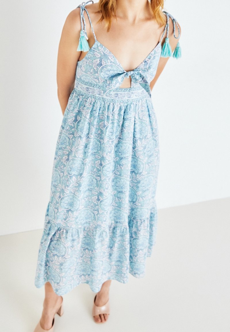 Blå klänning med blommönster, justerbara band med fransar på axelremmarna, lager-på-lager kjol och öppen ryggdesign. Slät tygtextur.
