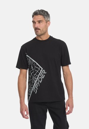 Homme aux cheveux gris courts portant un t-shirt noir à manches courtes avec un motif toile d'araignée blanche et le texte « SPYDER », debout devant un fond uni.
