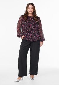 Frau mit langen braunen Haaren, die eine durchsichtige schwarze Bluse mit Blumenmuster und schwarze, weite Hosen trägt, posiert mit einer Hand in der Tasche und silbernen High Heels.