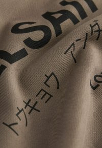 Un tissu brun avec une texture lisse présente un grand texte noir dans plusieurs langues. Le design comprend des lettres en gras et des couleurs contrastantes.
