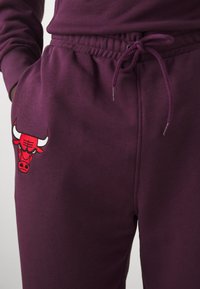 Pantaloni della tuta color marsala con vita elastica e cordoncino, caratterizzati da un logo di un toro ricamato in rosso sull'area della tasca sinistra. Texture morbida.