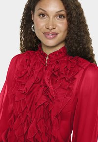 Piros blúz fodrozott elülső díszítéssel, magas nyakkal és gombos záródással. Áttetsző anyag lágy tapintással. Egyszerű kerek fülbevalók láthatók.