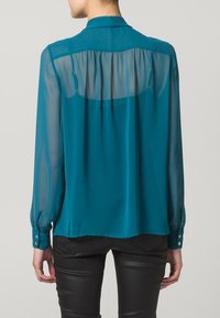 Blusa turchese trasparente con maniche lunghe e dettaglio della piega sul retro. Presenta un colletto e polsini con bottoni. Tessuto liscio con una texture leggera.
