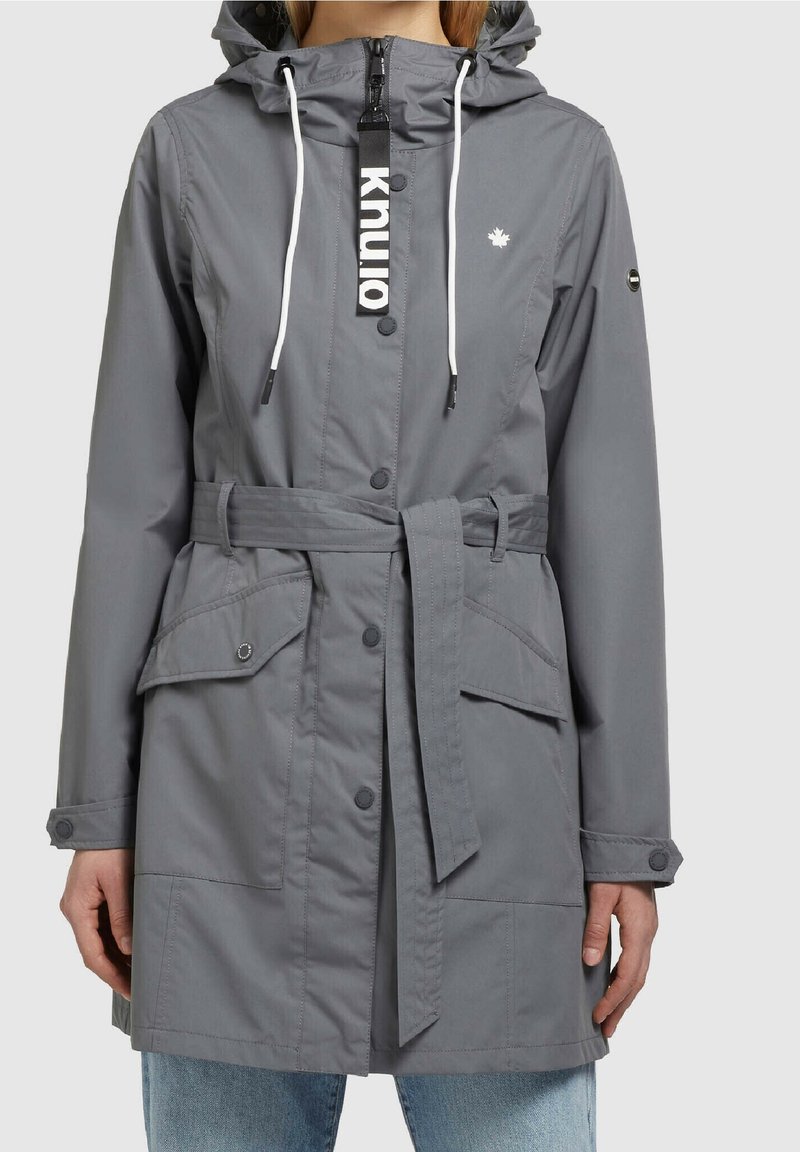 Imperméable gris à capuche avec cordons blancs, boutons à l'avant, ceinture à la taille, poches à rabat et petit logo blanc en forme de feuille d'érable sur la poitrine.