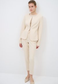 Beige tailored blazer and matching fitted trousers, made of structured fabric. Notable details include a round neckline and slim silhouette.