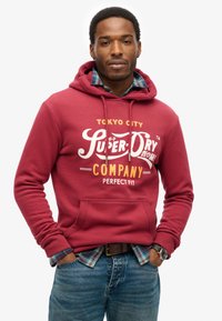 Rode sweatshirt met capuchon met de tekst "Super Dry Company" in wit en geel. Opvallende zak aan de voorkant, gedragen over een geruite shirt.