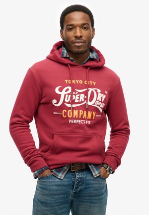 Sudadera roja con capucha que presenta el texto "Super Dry Company" en blanco y amarillo. Destaca un bolsillo en la parte frontal, llevada sobre una camisa de cuadros.