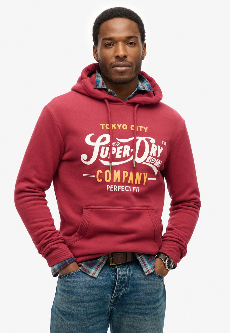 Rode sweatshirt met capuchon met de tekst "Super Dry Company" in wit en geel. Opvallende zak aan de voorkant, gedragen over een geruite shirt.