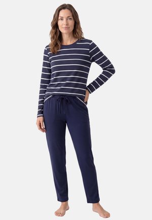 Donna in piedi a piedi nudi che indossa pantaloni blu navy e una maglia a maniche lunghe blu navy con strisce orizzontali bianche su sfondo bianco.