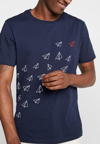 Marineblå bomull T-skjorte med et design av hvite papirfly, med ett rødt papirfly plassert på den nedre halvdelen.